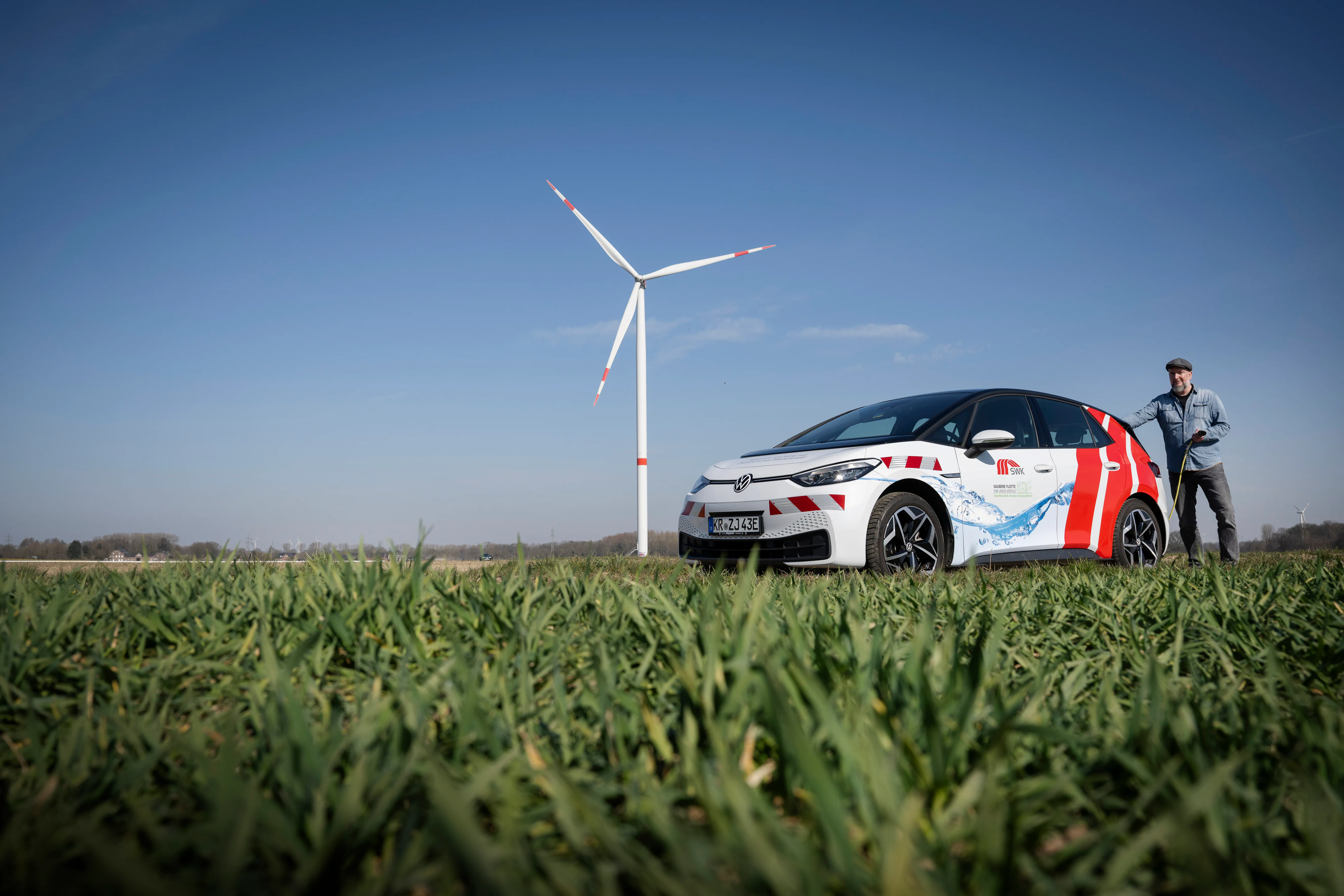 SWK ENERGIE E-Auto mit männlicher Person und Ladekabel auf einem grünen Feld mit einem Windrad im Hintergrund