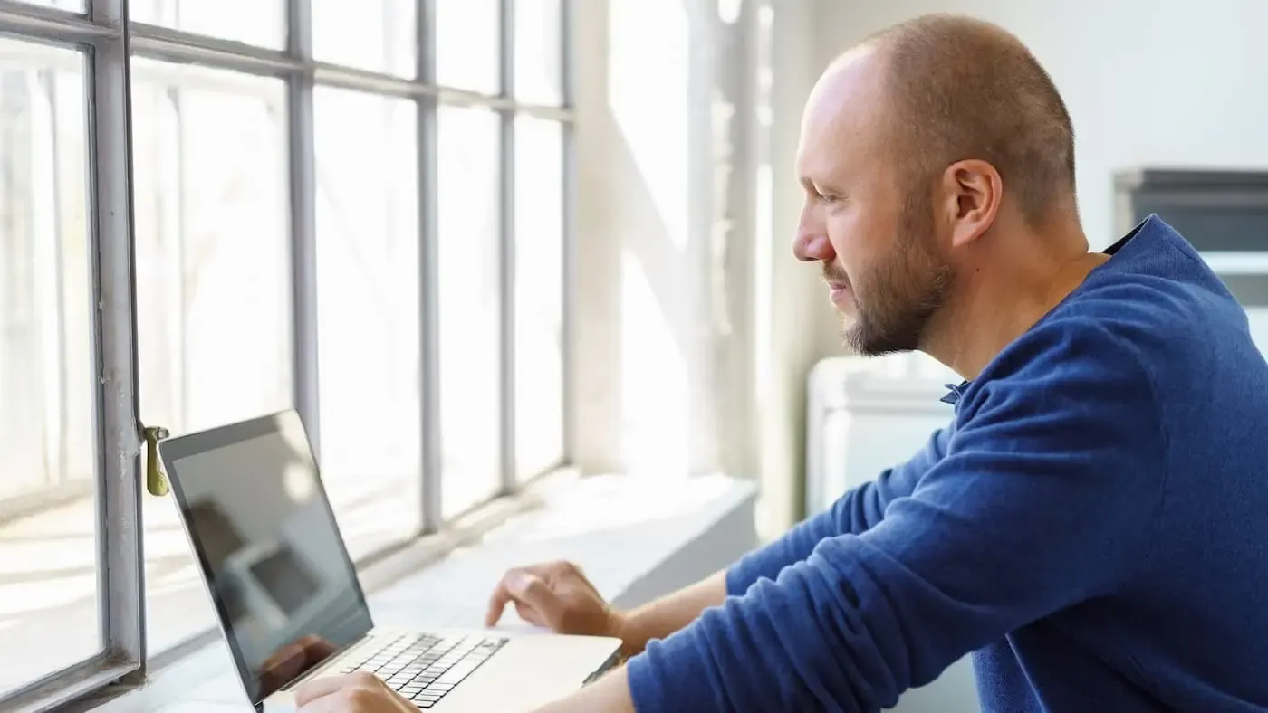 NGN Kollege steht mit Laptop auf der Fensterbank am Fenster