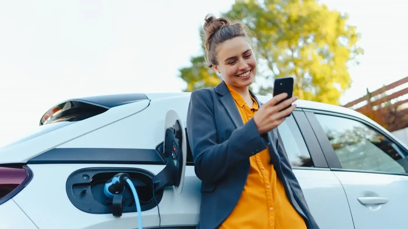 Frau steht am E-Auto und schaut aufs Handy